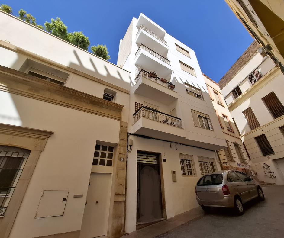 Edificio de apartamentos blanco con balcones, coche aparcado en la calle. Día soleado.