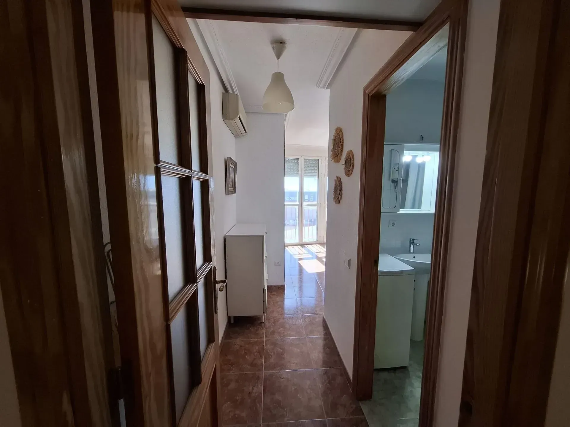 Pasillo con puertas abiertas. Suelo de baldosas marrones, paredes blancas y luz solar brillante al fondo.