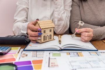 Dos personas en un escritorio, una sosteniendo una maqueta de casa, la otra una llave, al lado de un cuaderno y planos.