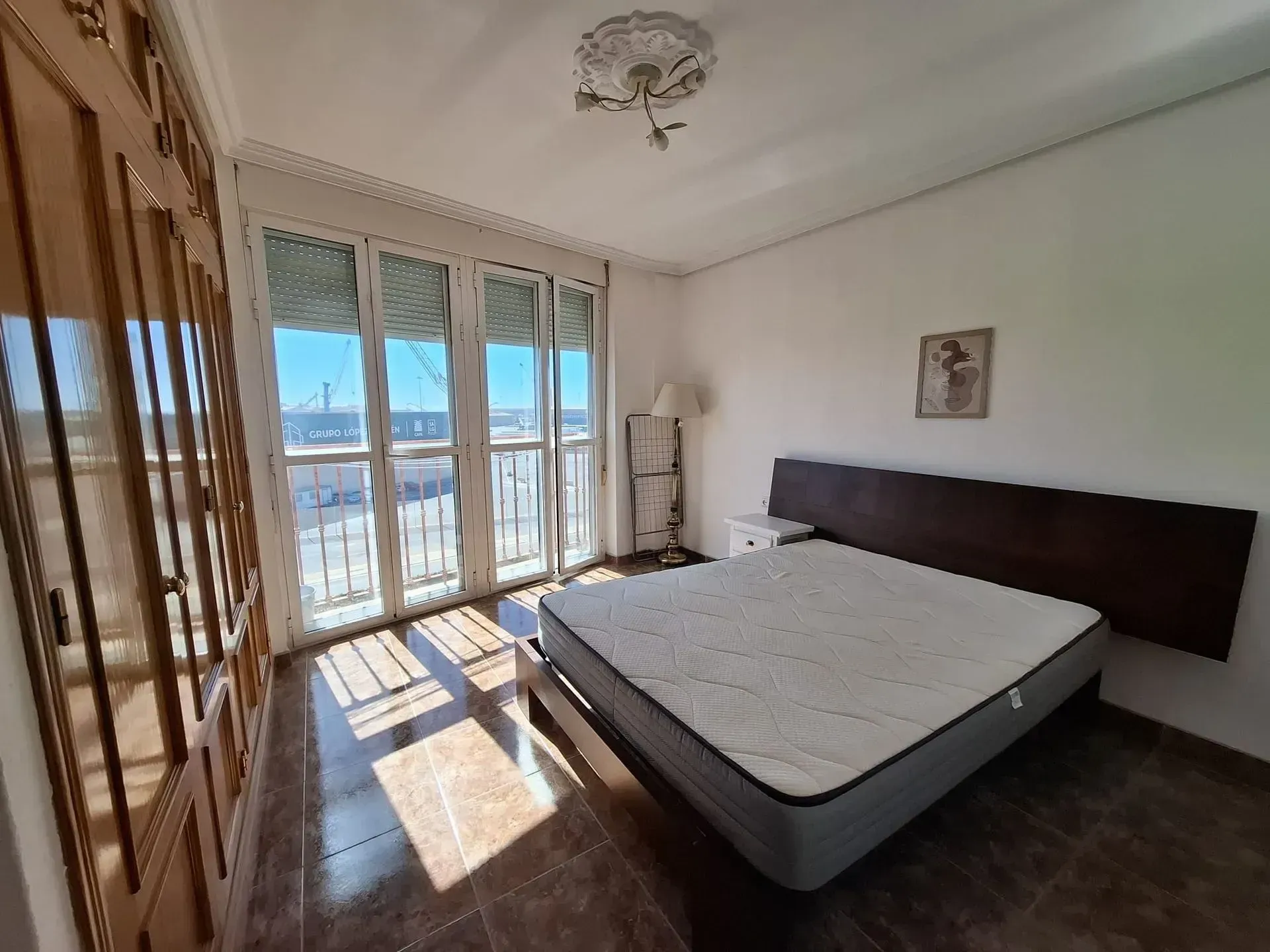 Dormitorio con cama, armario y puertas de balcón; entra la luz del sol.