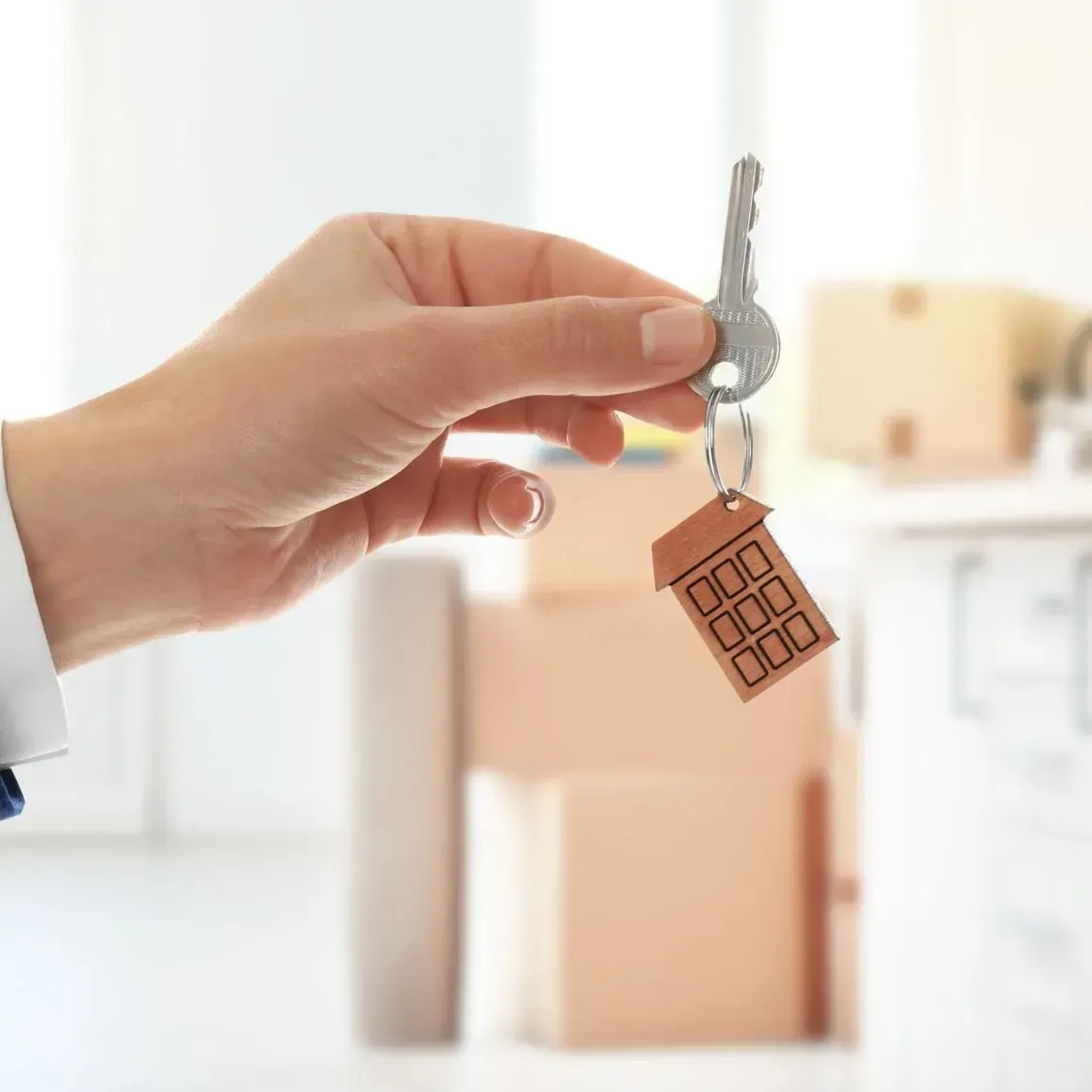 Mano sosteniendo la llave de la casa con llavero en forma de casa, cajas en el fondo, interior claro.