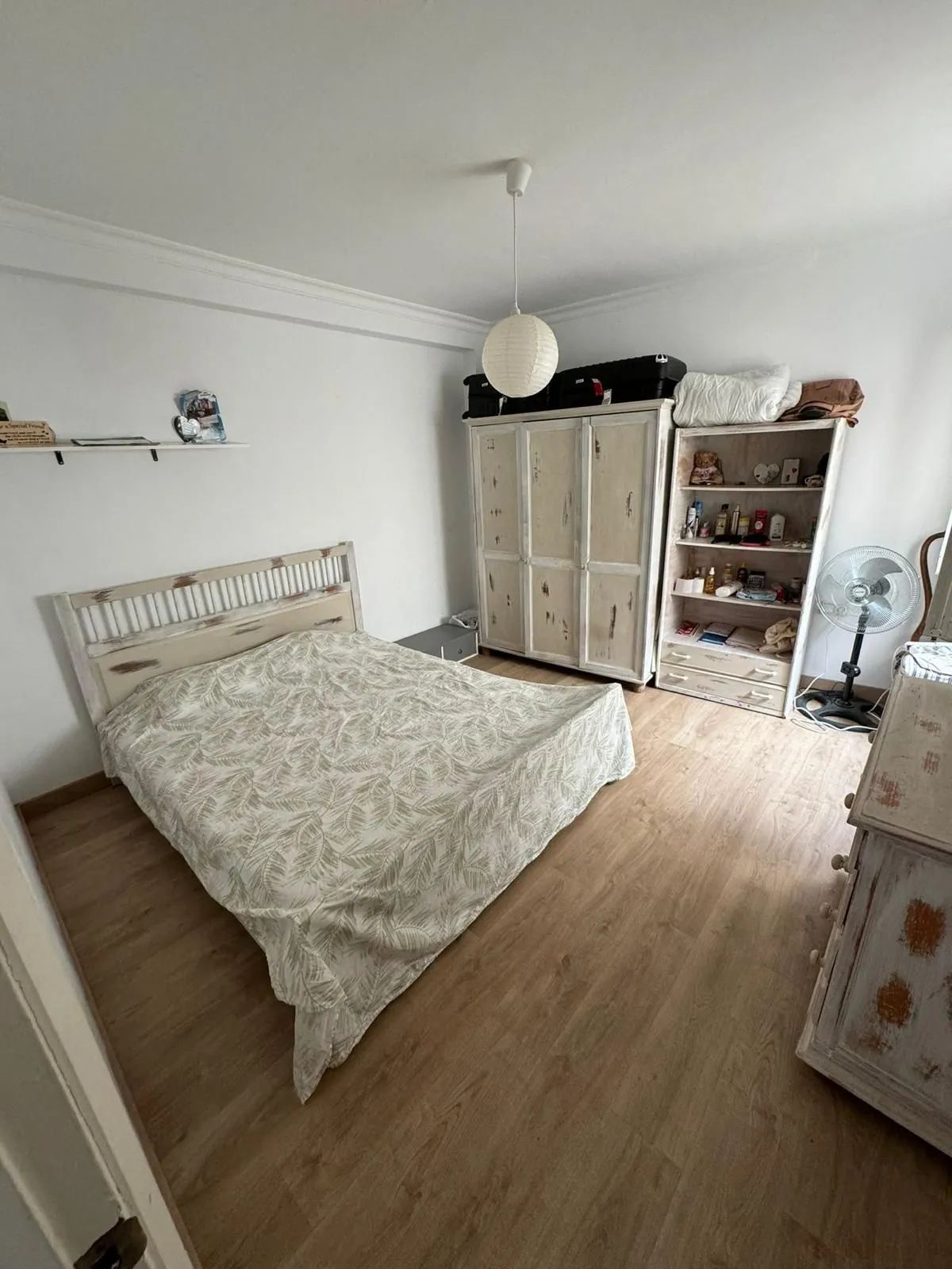 Dormitorio con cama, armario, estantería y ventilador sobre suelo de madera.