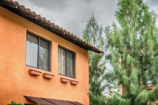 Edificio de estuco naranja con ventanas oscuras, techo marrón y jardineras; altos árboles verdes en el fondo.