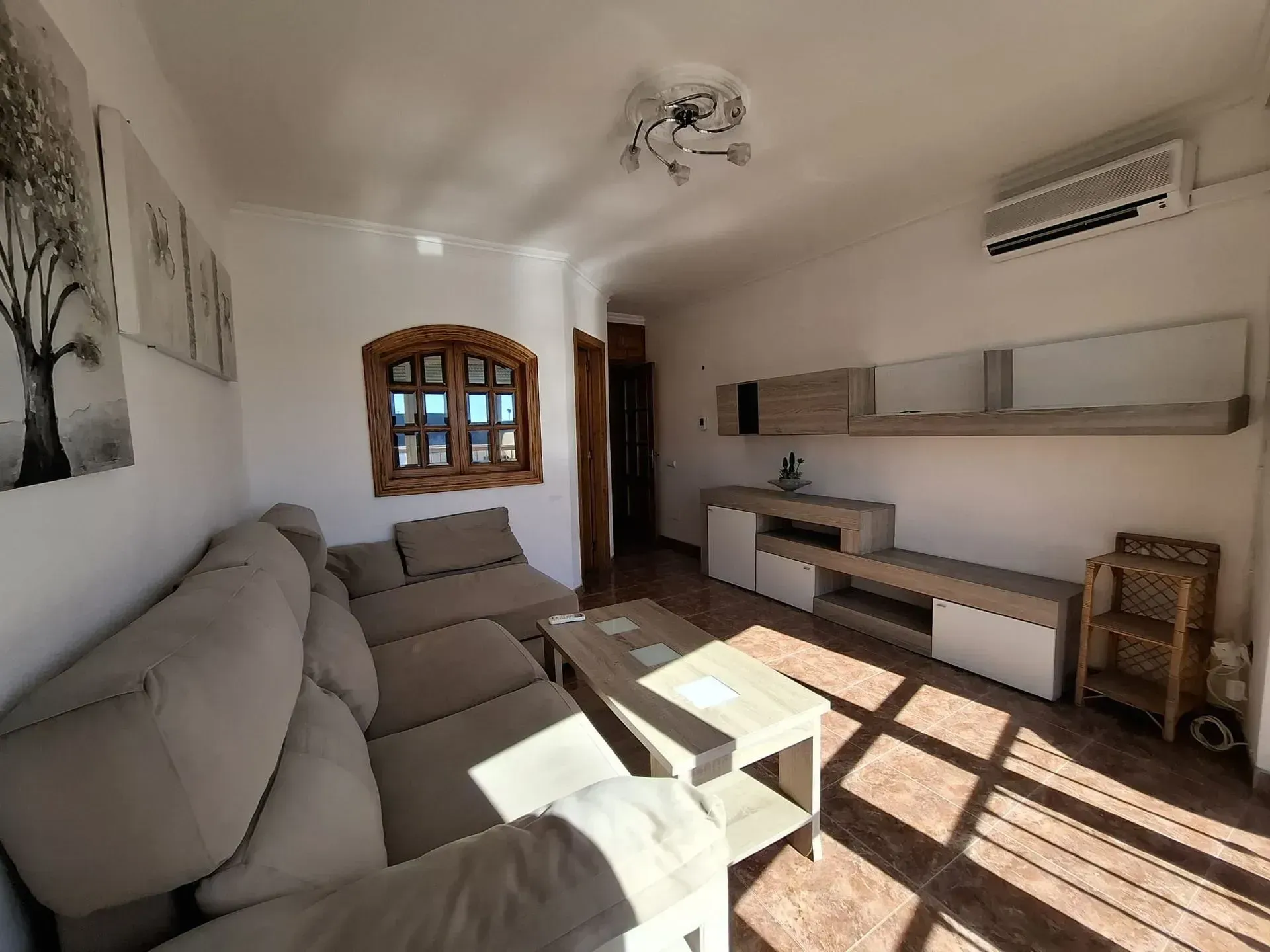 Sala de estar con sofá seccional color tostado, ventana de madera y centro de entretenimiento incorporado; luz solar.