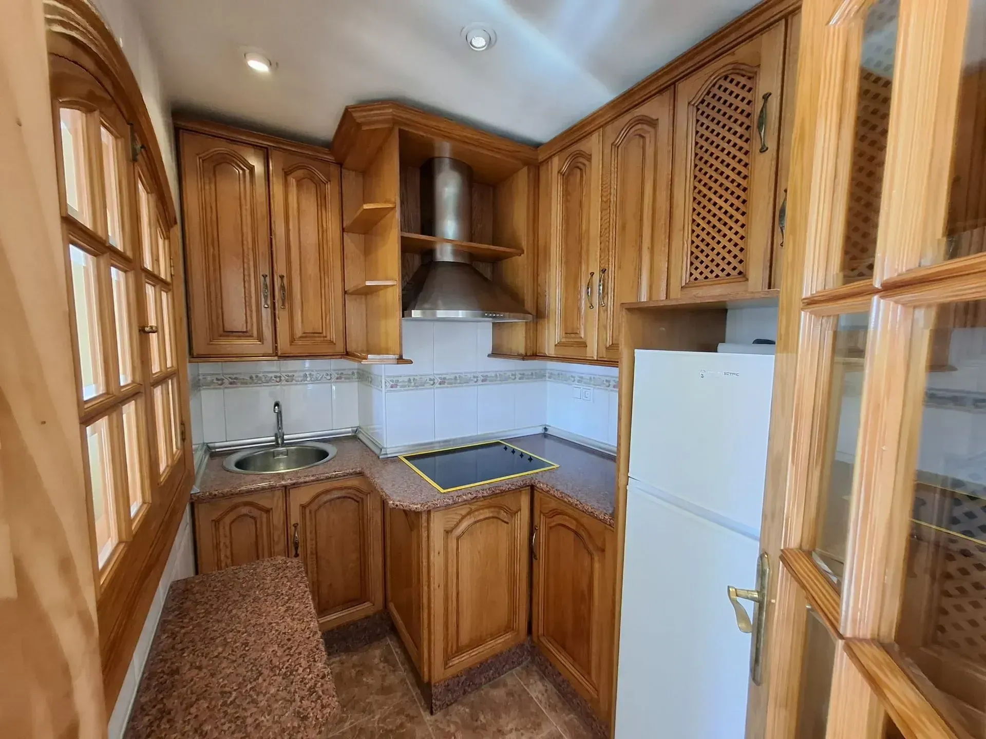 Cocina pequeña con gabinetes de madera, fregadero, estufa y refrigerador. Tonos marrones y blancos.