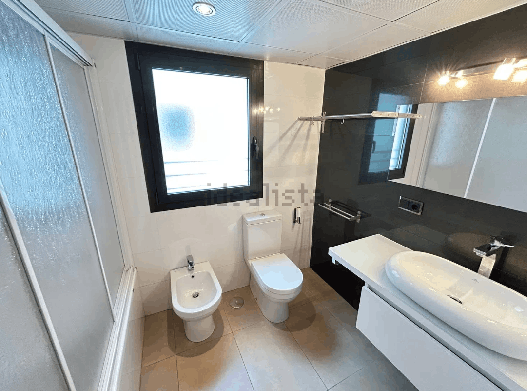 Baño moderno con inodoro, bidé, lavabo, ventana y cabina de ducha. Presenta azulejos blancos y negros.