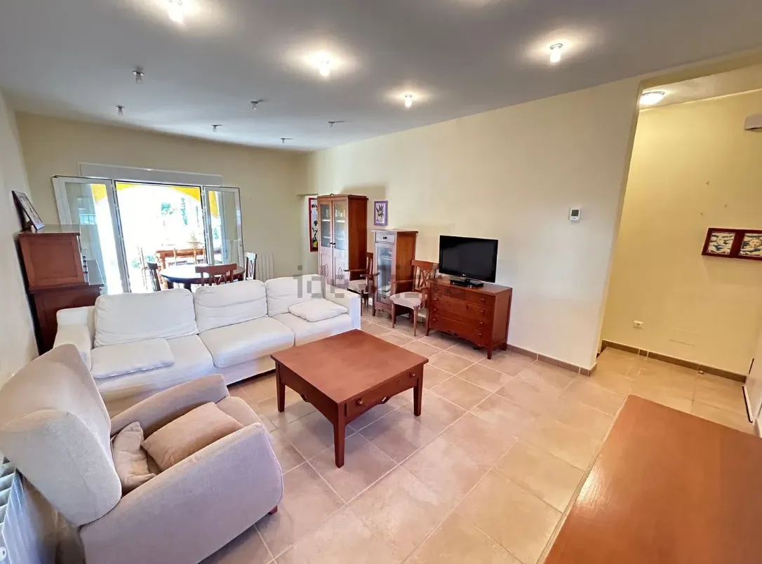 Sala de estar con sofá blanco, muebles de madera, TV y acceso a un comedor exterior.