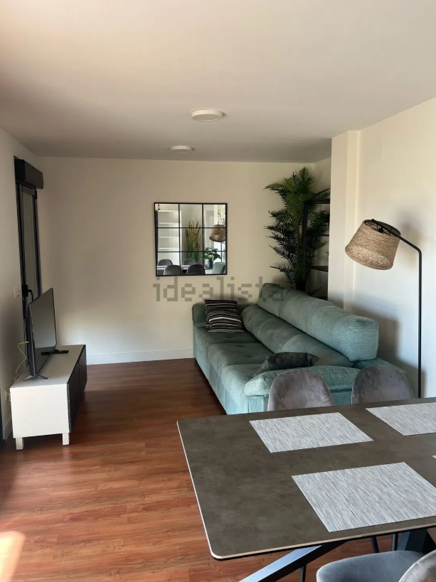 Sala de estar con sofá verde azulado, espejo, televisión y mesa de comedor sobre piso de madera.