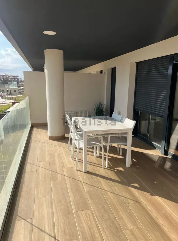 Balcón con mesa y sillas blancas y puerta con persianas. Suelo de madera, altas columnas blancas y barandilla de cristal.