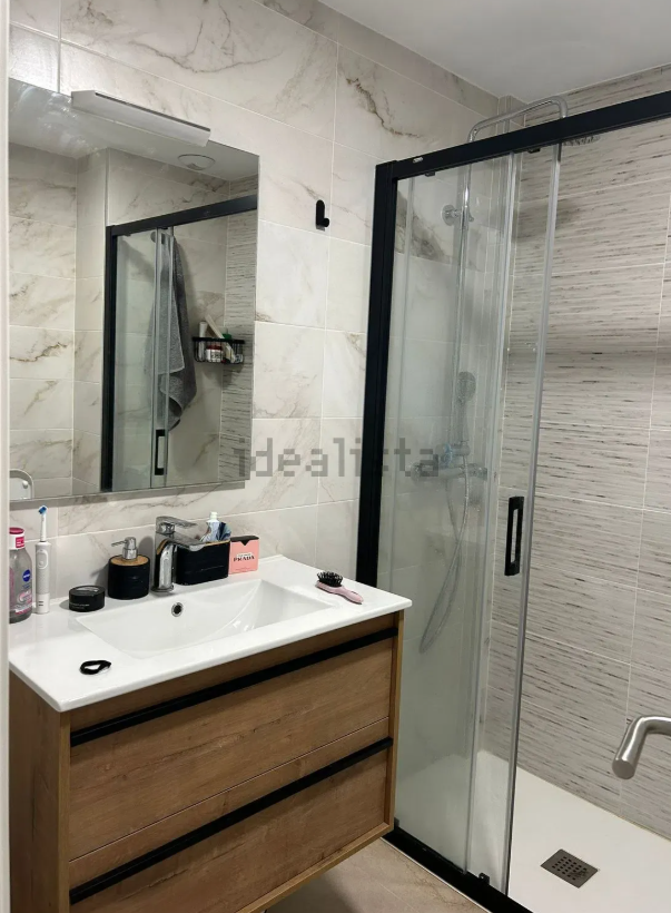 Baño con tocador de madera, lavabo blanco, espejo con marco y mampara de ducha de cristal.