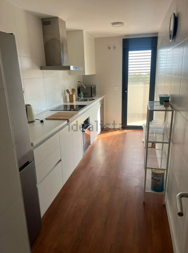 Cocina moderna con gabinetes blancos, electrodomésticos de acero inoxidable y pisos de madera. Una puerta da a un balcón.