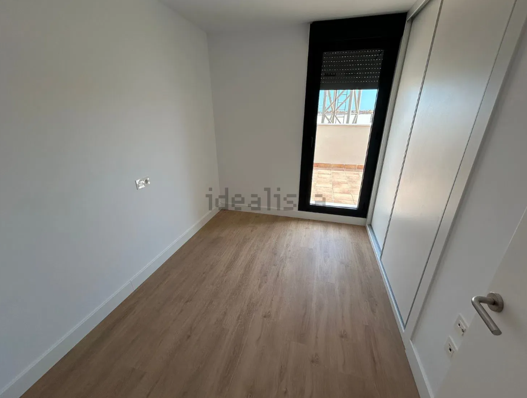 Habitación pequeña y vacía con suelo de imitación madera clara, paredes blancas y una ventana con molduras negras.