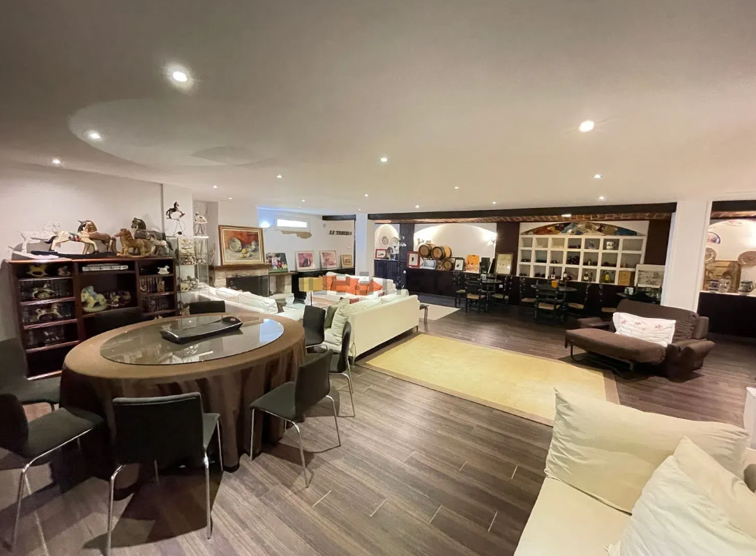 Amplia sala de estar con mesa de comedor ovalada, sofás, estanterías con decoración y chimenea. Suelo oscuro.