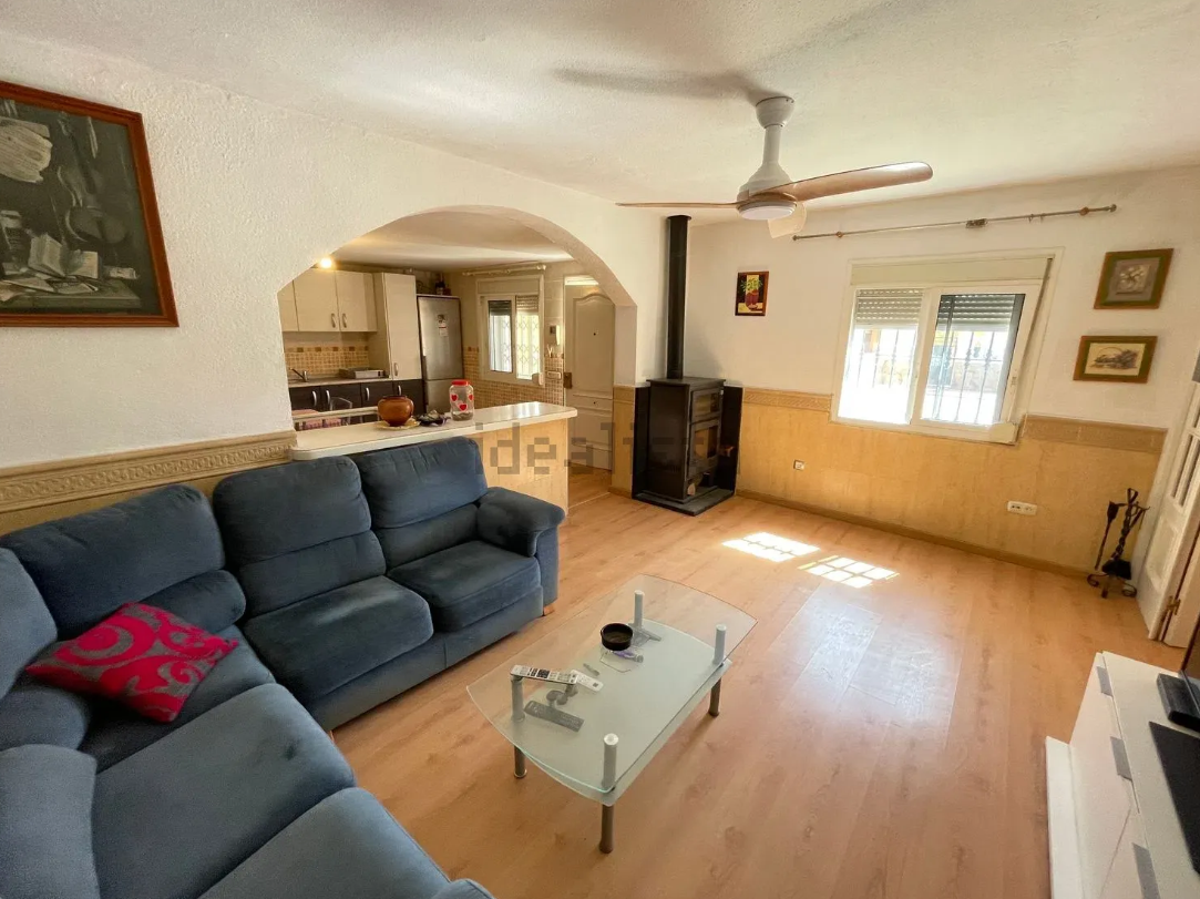 Sala de estar con sofá azul, puerta arqueada a la cocina y estufa de leña.