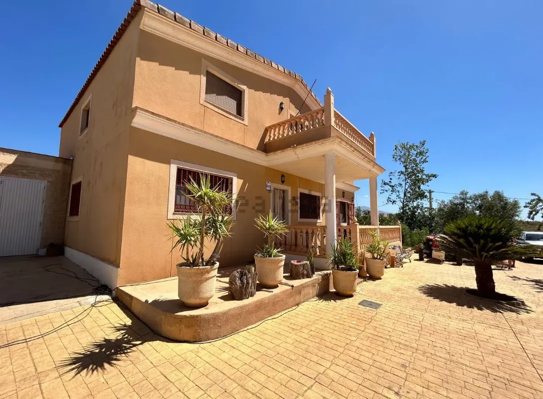 Casa de dos pisos con exterior de estuco color canela, pequeño balcón y plantas en macetas en un día soleado.