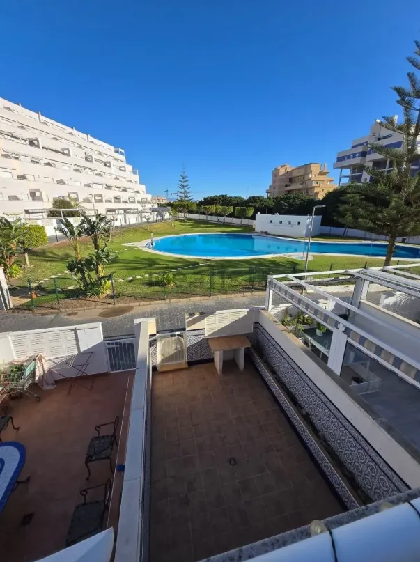 Vista desde un patio hacia una piscina, césped y edificios bajo un cielo azul.