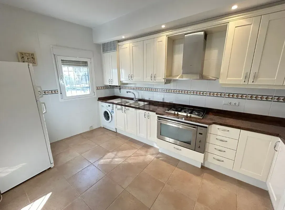 Cocina color crema con gabinetes, estufa y refrigerador. Encimeras marrones y piso de baldosas. Luz natural por una ventana.