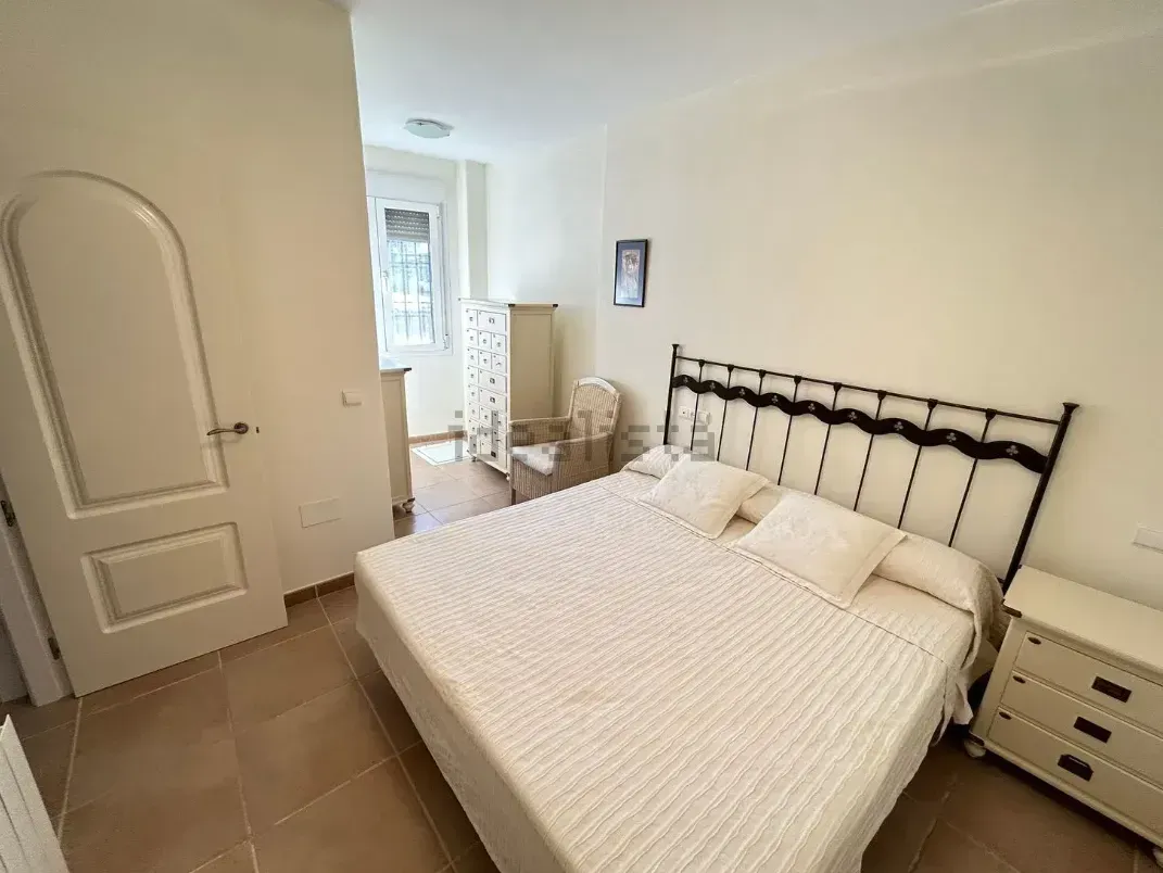 Dormitorio con cama, cómoda y puerta. Paredes color crema, suelo de baldosas y una ventana al fondo.