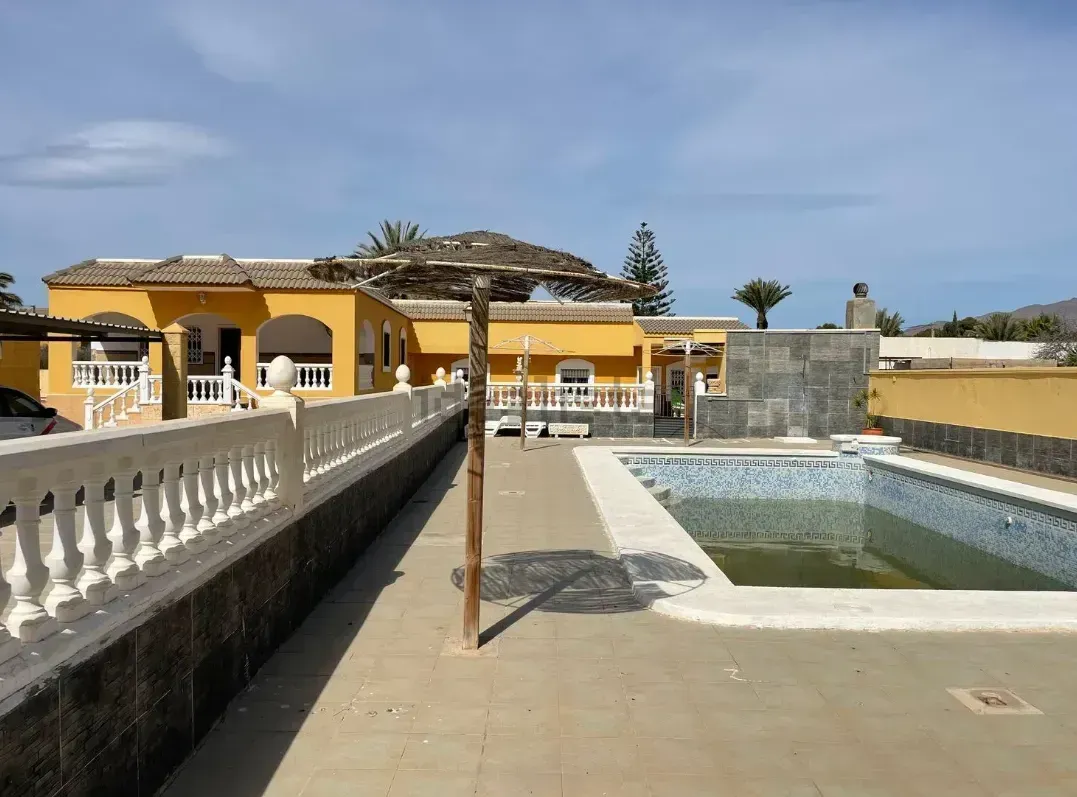 Villa amarilla con piscina drenada, balaustrada blanca y cielo azul.