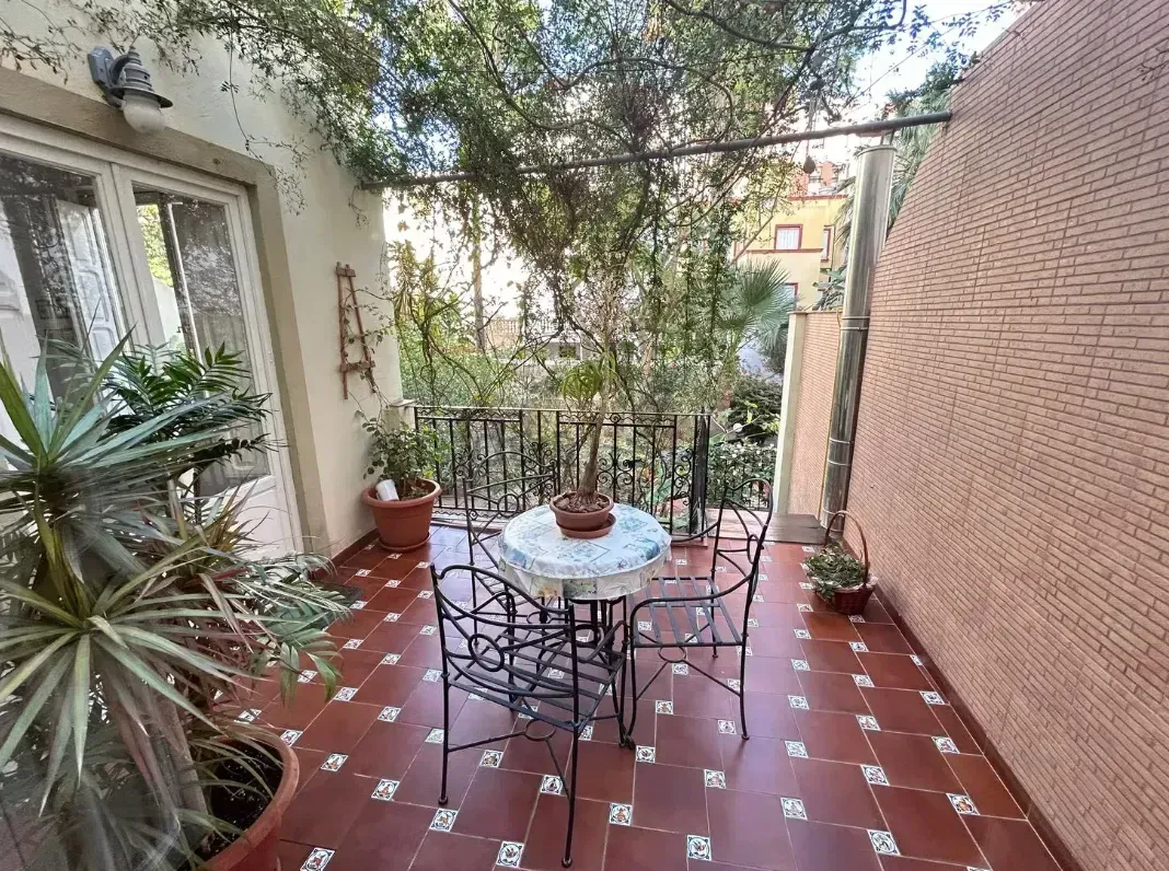 Pequeño patio con mesa y sillas, piso de baldosas rojas, plantas y una vista parcial de un edificio.