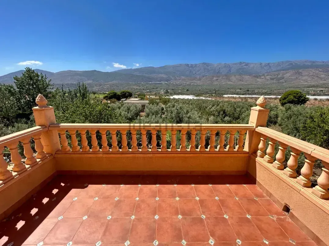 Terraza con balaustrada naranja, suelo de baldosas rojas, con vistas a un valle y montañas bajo un cielo azul.