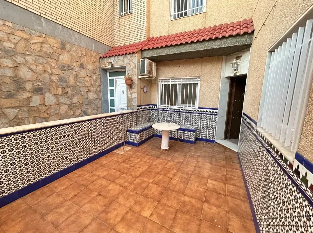 Pequeño patio con paredes de baldosas, suelo marrón, mesa blanca y entrada a un edificio.