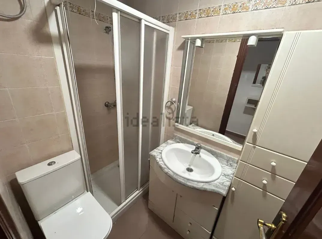 Baño pequeño con inodoro, ducha, tocador y espejo. Tonos blanco y beige.