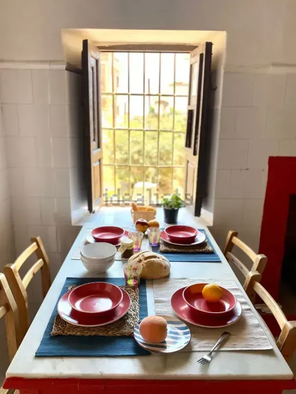 Mesa de desayuno para dos frente a una ventana. Platos rojos, manteles individuales azules, pan y fruta.