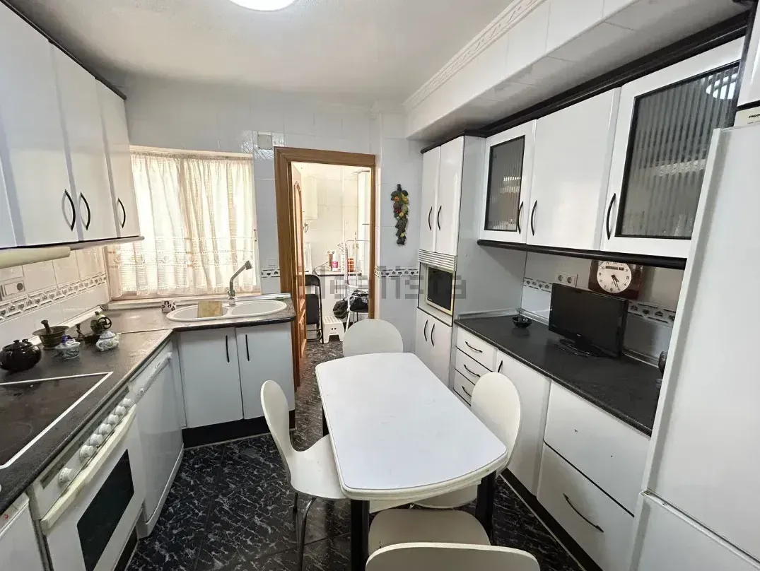 Cocina blanca con gabinetes, estufa, fregadero, mesa, sillas y puerta.