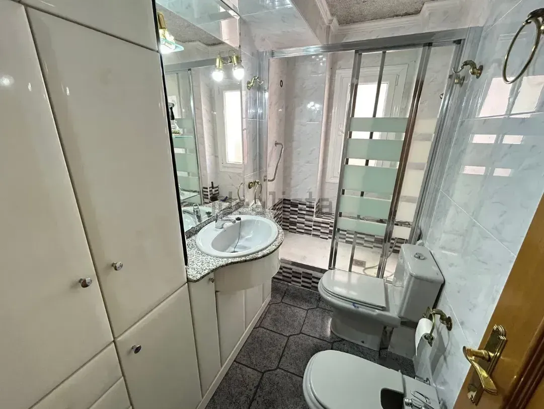 Interior de baño con muebles blancos, lavabo, inodoro y ducha. Suelo de baldosas negras y grises.