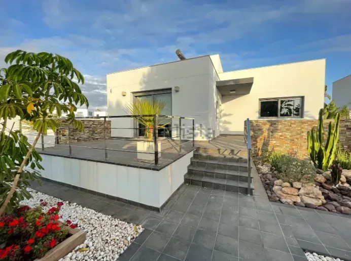 Casa blanca moderna con patio, jardín y escaleras que conducen hacia arriba; cielo azul brillante.