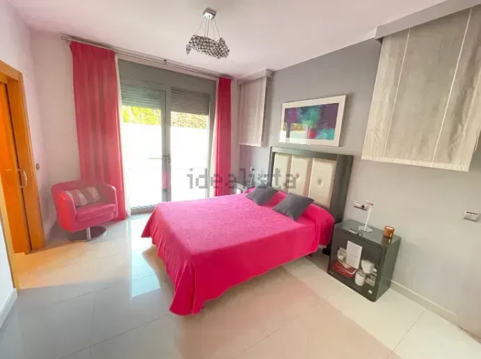 Dormitorio con colcha, cortinas y sillón de color rosa; paredes grises, suelo blanco y una puerta que da al exterior.