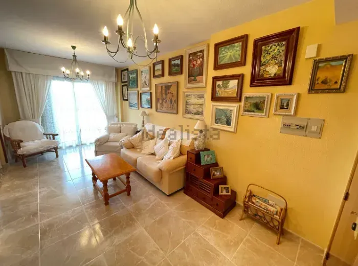 Sala de estar con paredes de colores claros, sofá, muchos cuadros enmarcados y una lámpara de araña; luz desde un balcón.