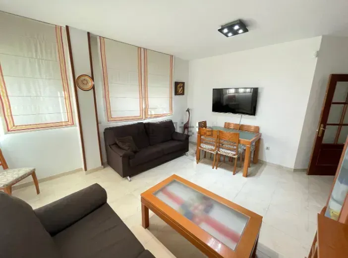 Sala de estar con sofá marrón, mesa de comedor de madera y TV en una pared blanca.