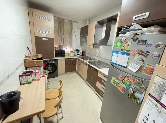 Cocina pequeña con gabinetes en tonos madera, electrodomésticos y una mesa con sillas. Refrigerador cubierto de imanes.