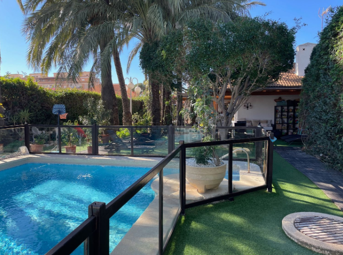 Área de piscina con cerca de vidrio, césped verde y árboles bajo un cielo soleado.