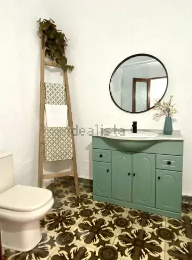 Baño con tocador verde, espejo redondo, piso de baldosas florales y una escalera de madera con toallas.