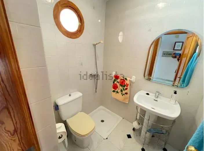 Pequeño baño con azulejos blancos, inodoro, ducha, lavabo, espejo y toallero.