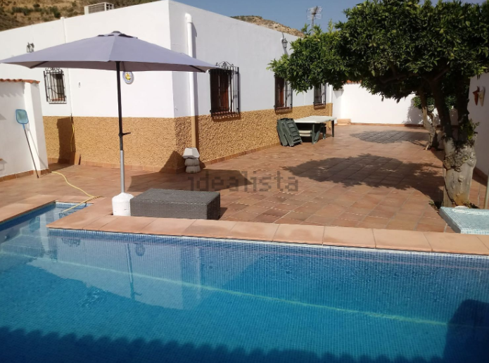 Casa con piscina y patio. El edificio es blanco y tostado, con una sombrilla gris sobre una caja de mimbre oscura.