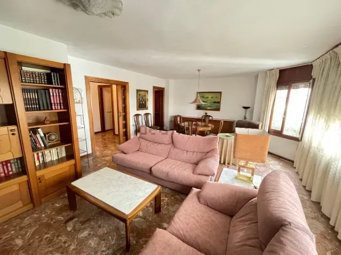 Sala de estar con sofás rosas, estantería de madera y mesa de comedor, con luz solar desde una ventana.