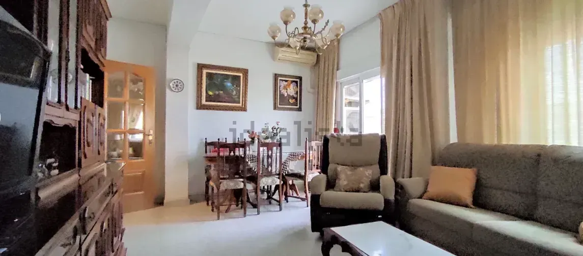 Sala de estar con sofá, sillón, juego de comedor, cuadros y lámpara de araña. Cortinas beige y mesa de centro blanca.