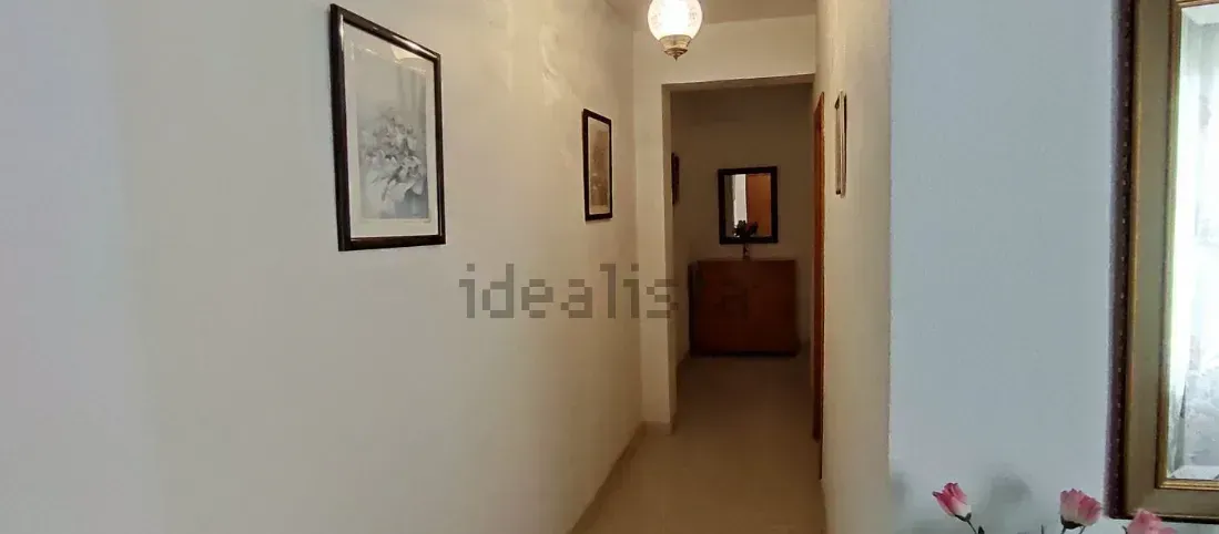 Pasillo estrecho con paredes blancas, obras de arte enmarcadas y una lámpara colgante.