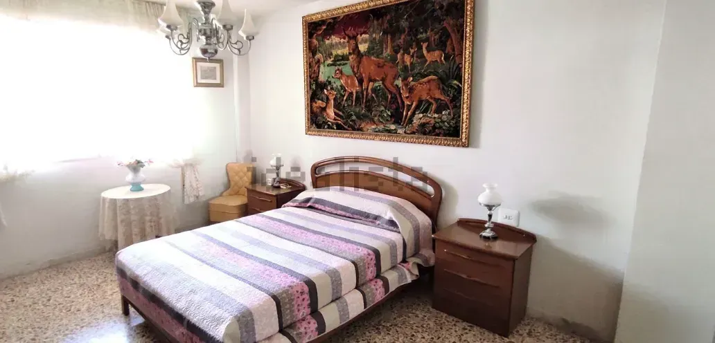 Dormitorio con cama, mesita de noche, tapiz y lámpara de araña. Ventana a la izquierda.