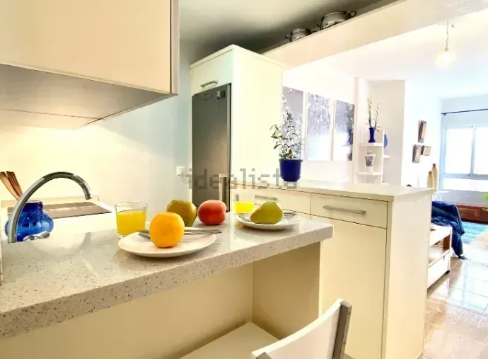 Encimera de cocina con frutero, abierta a zona de estar con muebles blancos y refrigerador.