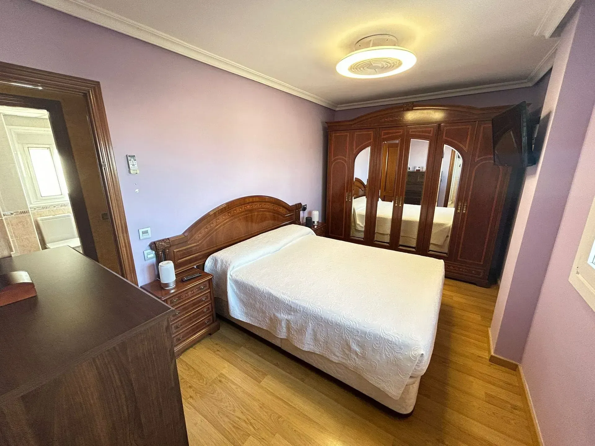 Dormitorio con muebles de madera, incluyendo cama, mesitas de noche y armario. Paredes color lavanda y suelo de madera clara.
