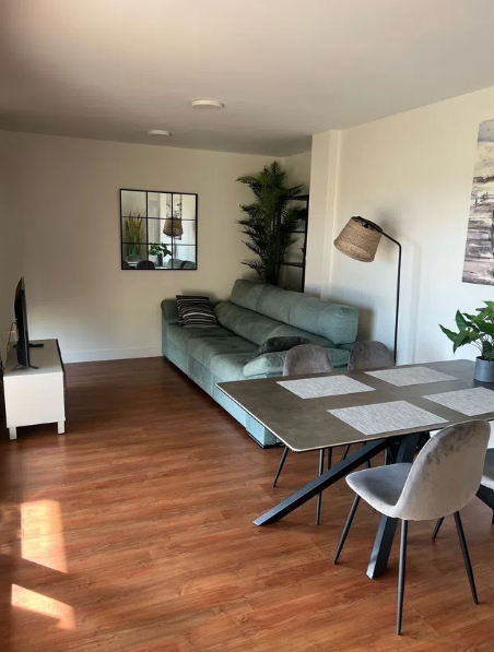 Sala de estar con sofá verde azulado, espejo, televisión y mesa de comedor sobre piso de madera.