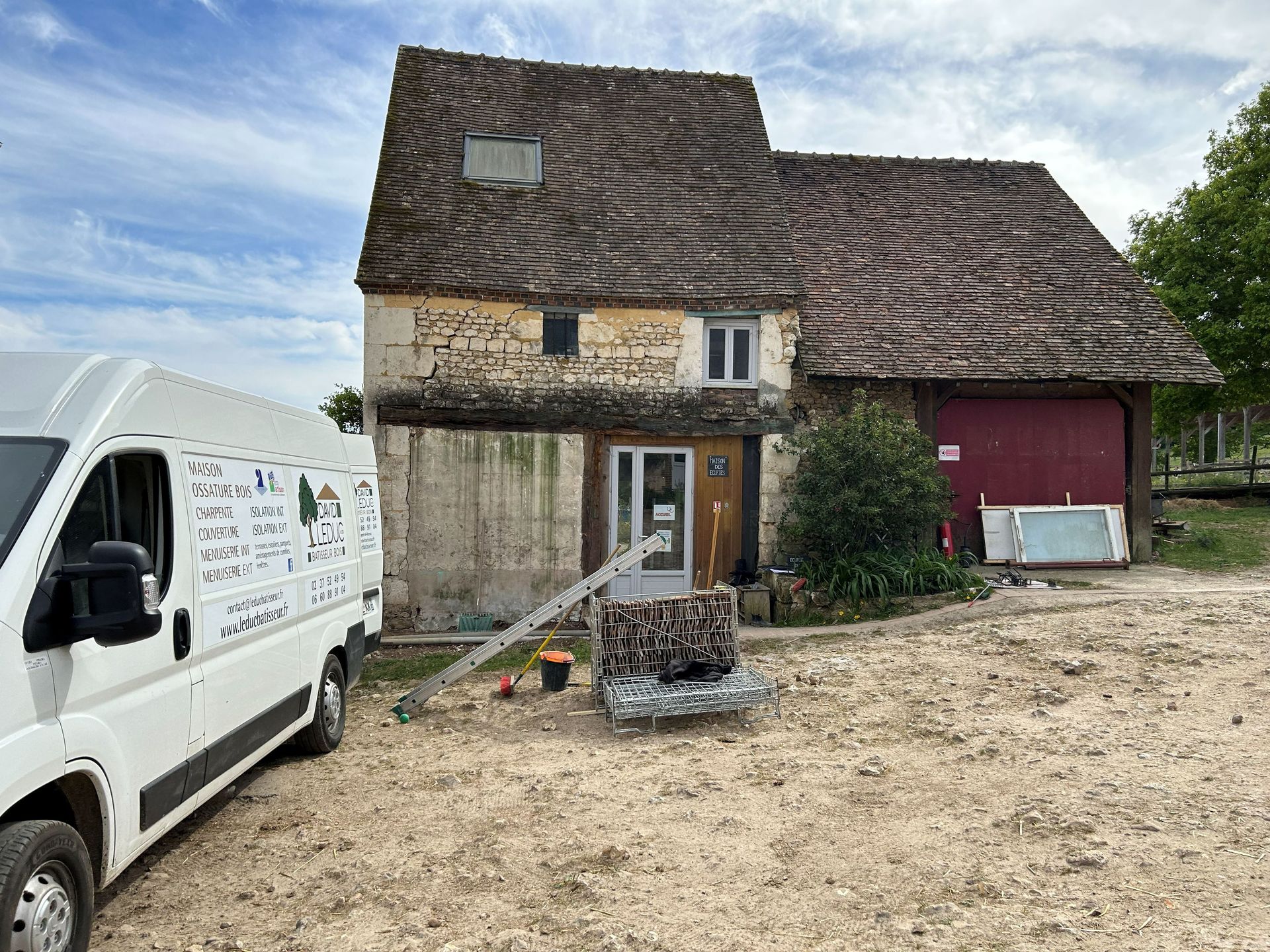 Maison rurale en pierre au toit délabré, camionnette blanche garée à gauche et cour de gravier sous un ciel nuageux