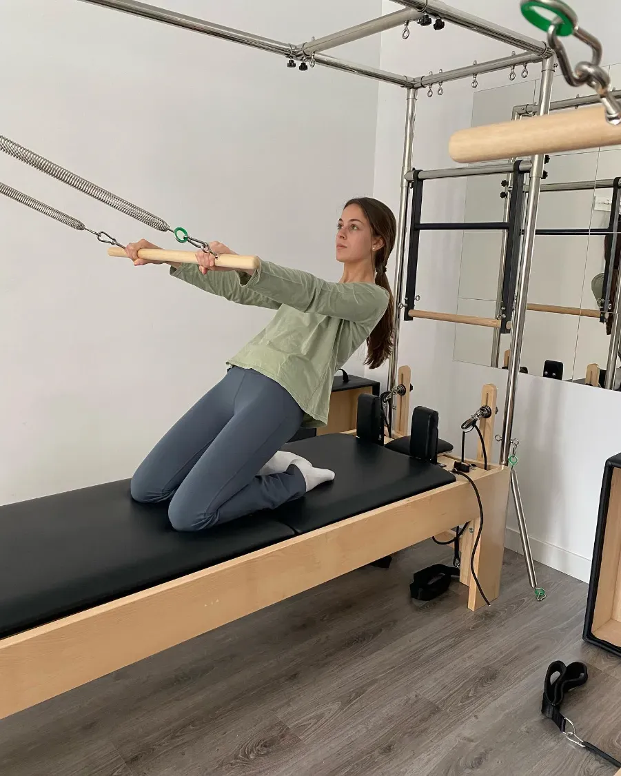Mujer arrodillada en una máquina de Pilates, sosteniendo manijas, haciendo ejercicio en una habitación luminosa.