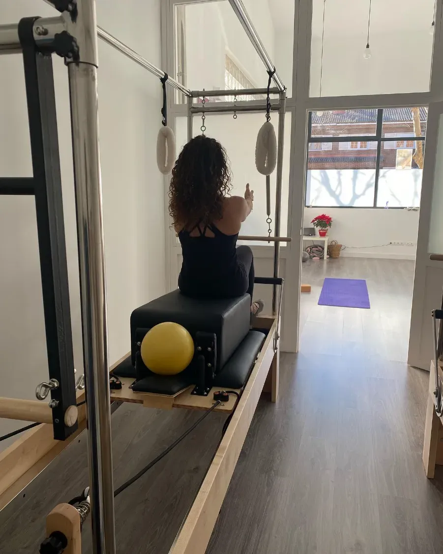 Mujer en una máquina de pilates, realizando ejercicios en un estudio luminoso. Equipo beige y negro, suelo de madera.