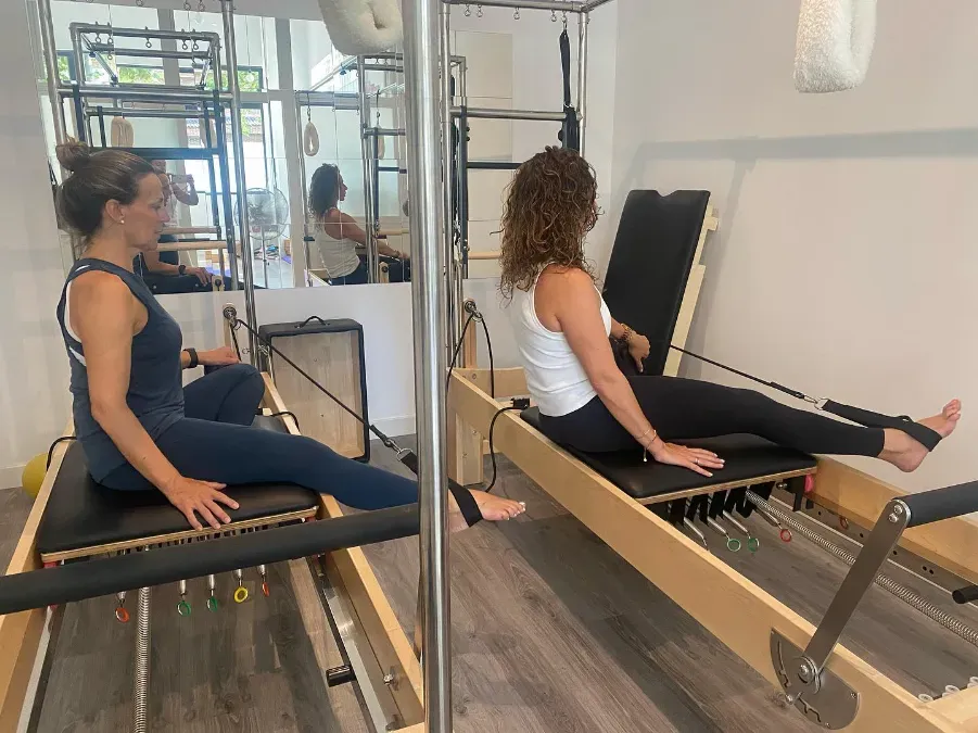 Dos personas haciendo ejercicio en máquinas de Pilates reformer en un estudio luminoso, una sentada y la otra reclinada usando correas.